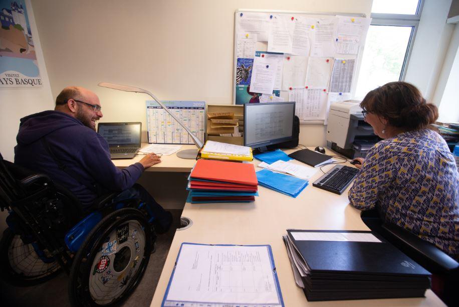 Un homme en fauteuil roulant et une femme discutent à leurs bureaux.